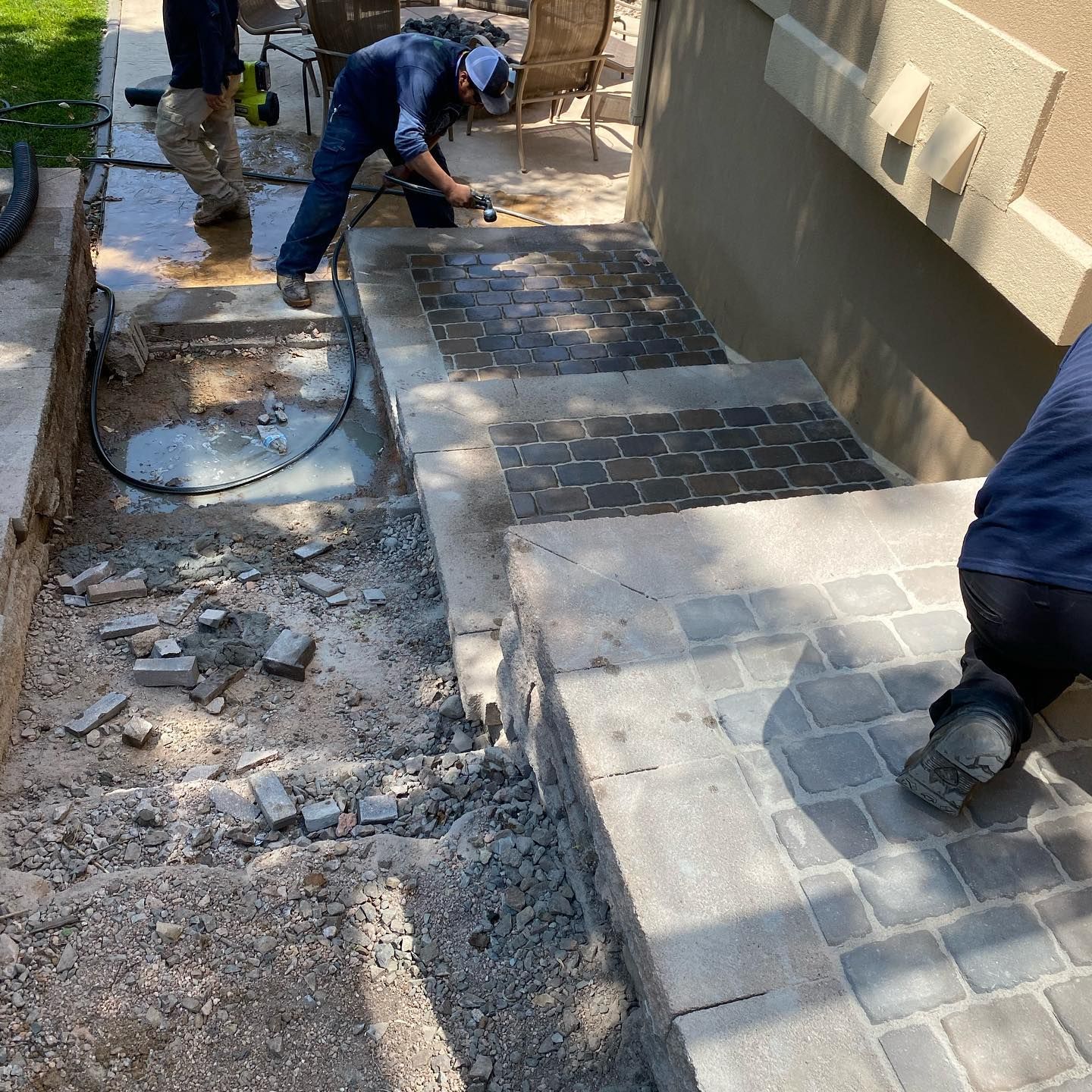 Construction workers laying pavers for steps. Gray and black pavers are being installed near a concrete wall.