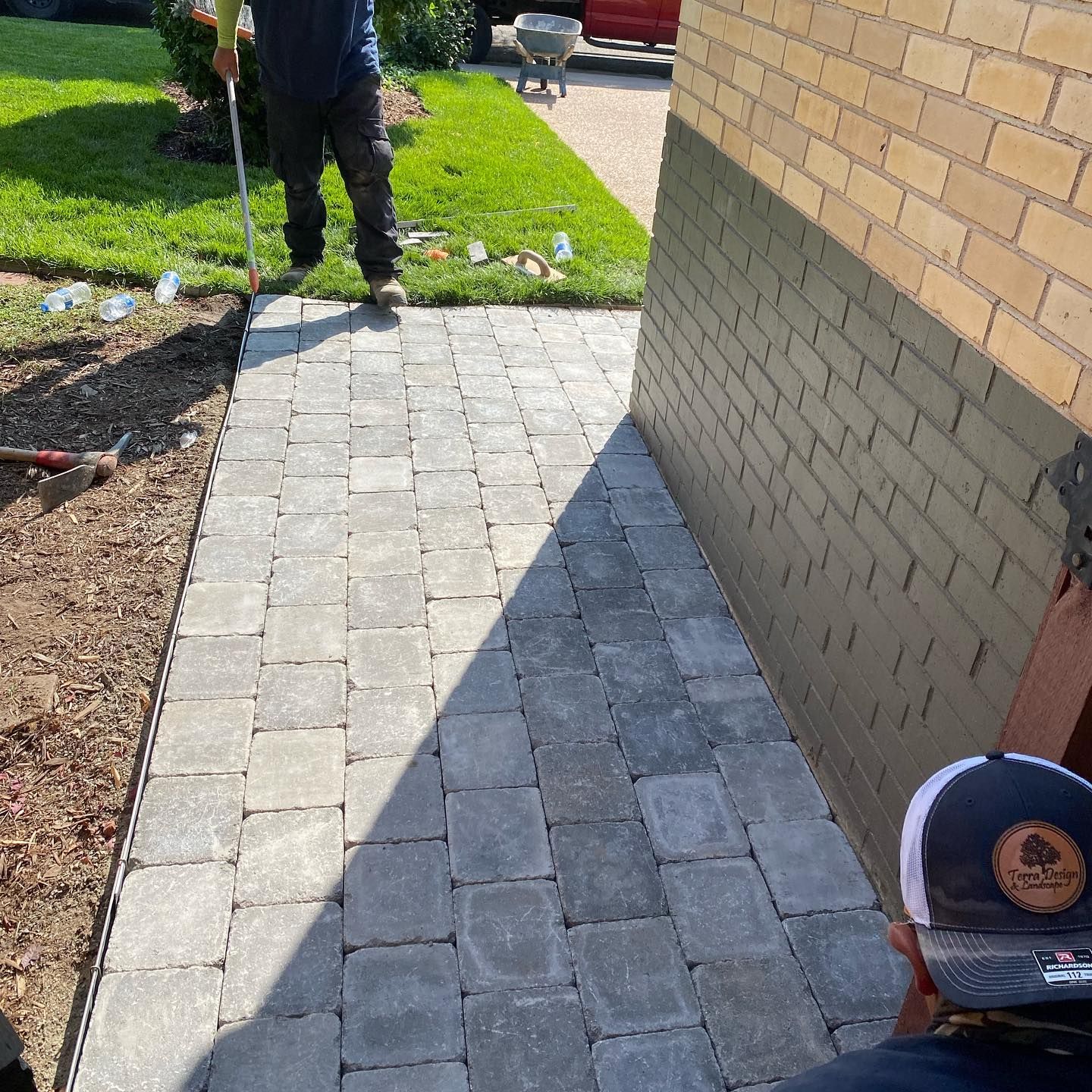 A person inspects a newly paved walkway made of gray pavers next to a brick wall; another person is present.