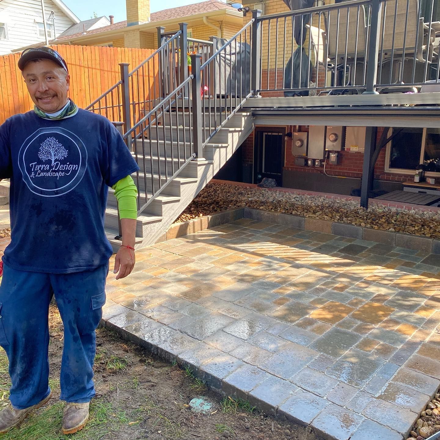 Man in blue shirt and jeans smiles, standing by a newly paved patio area under a deck with stairs.