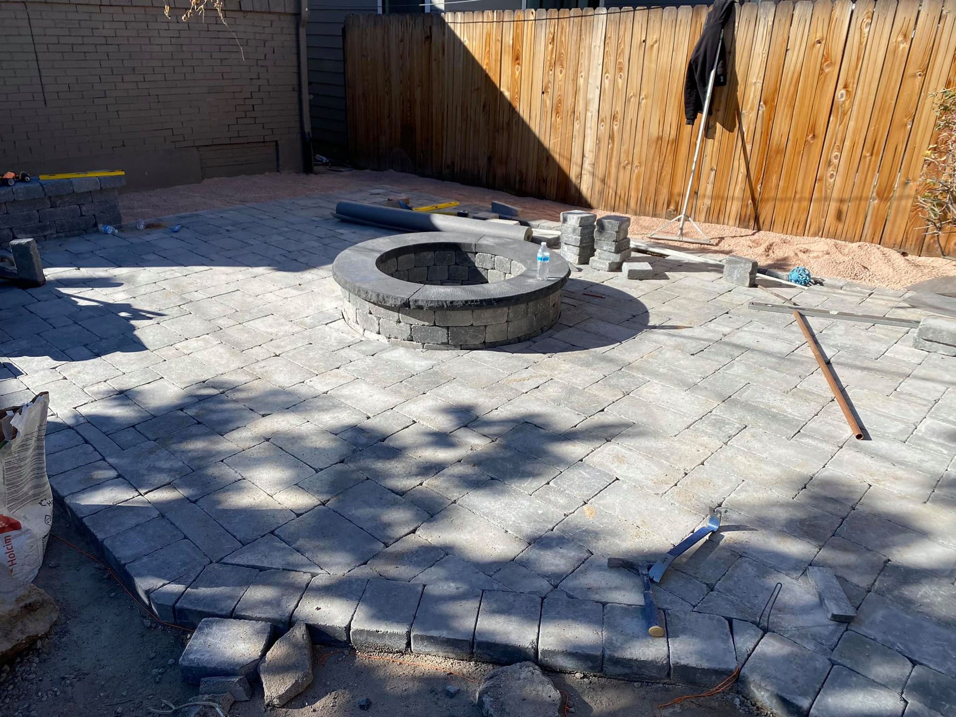 Backyard patio under construction with a stone fire pit in the center, surrounded by gray paving stones.