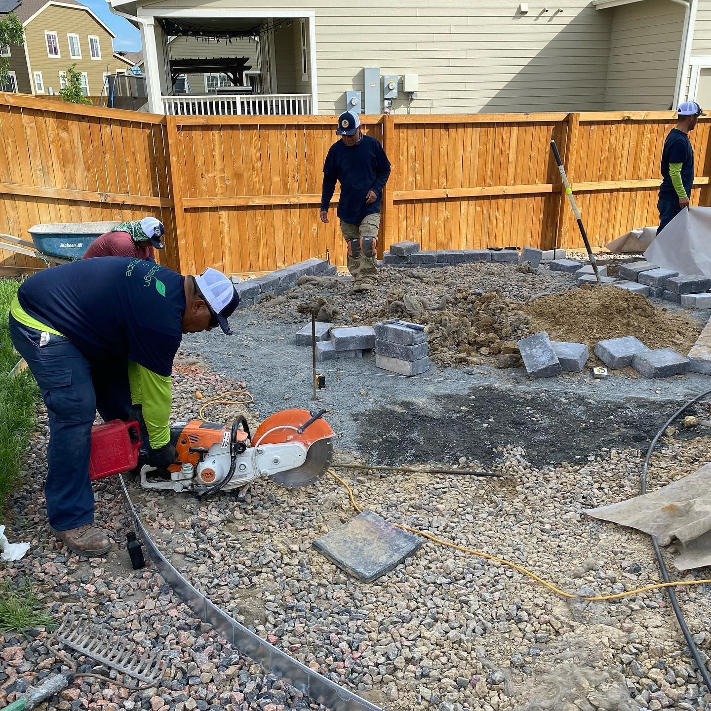 Landscapers cutting stone for a patio in a backyard with a wooden fence. Workers are wearing hats and safety gear.