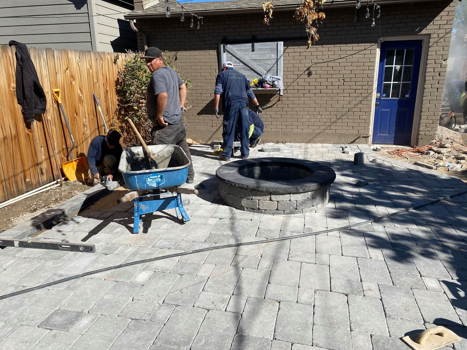 Workers install a brick patio with a fire pit in a backyard. The sky is clear, and a shed sits in the background.