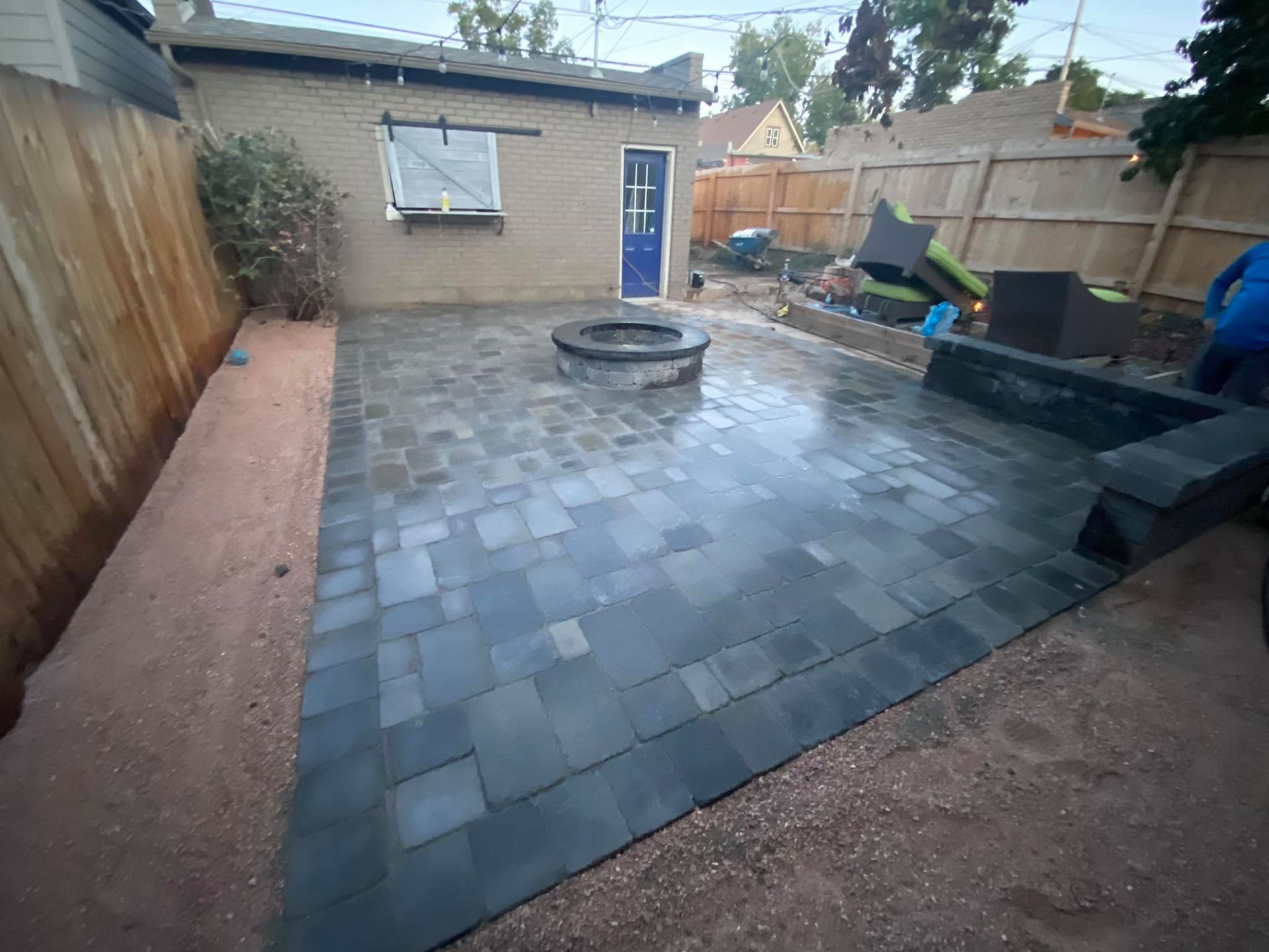 A backyard patio with dark gray pavers, a fire pit, and a retaining wall, backed by a tan stucco building with a blue door.