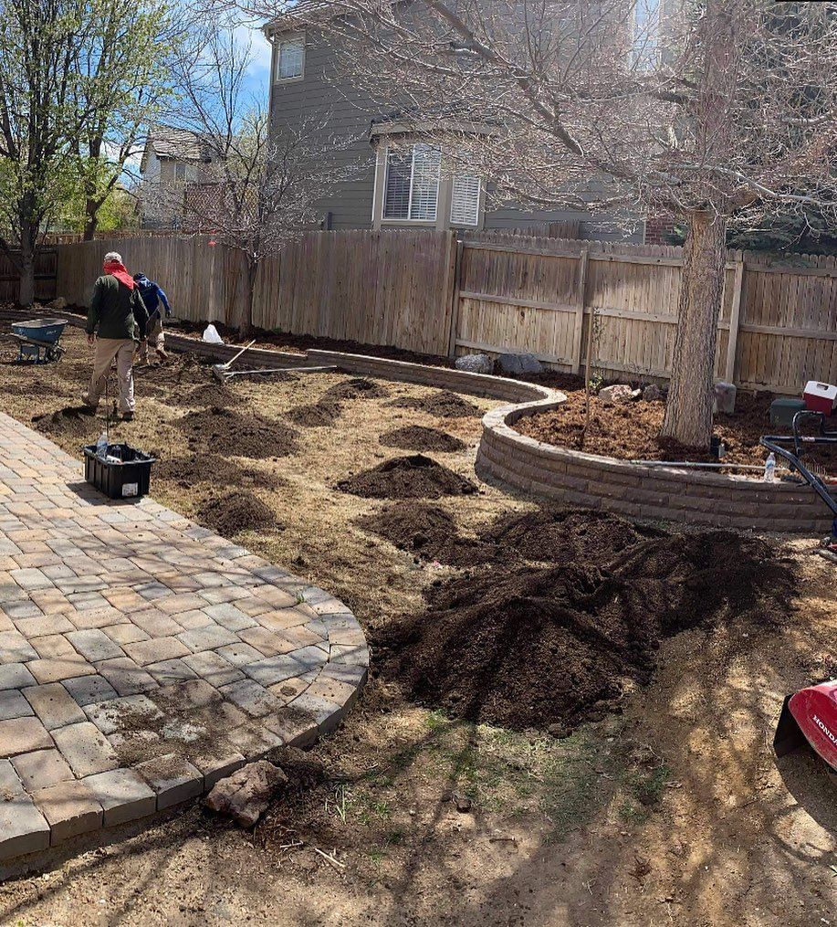 Backyard landscaping in progress. Two workers spread dirt, mulched beds, and brick patio visible.