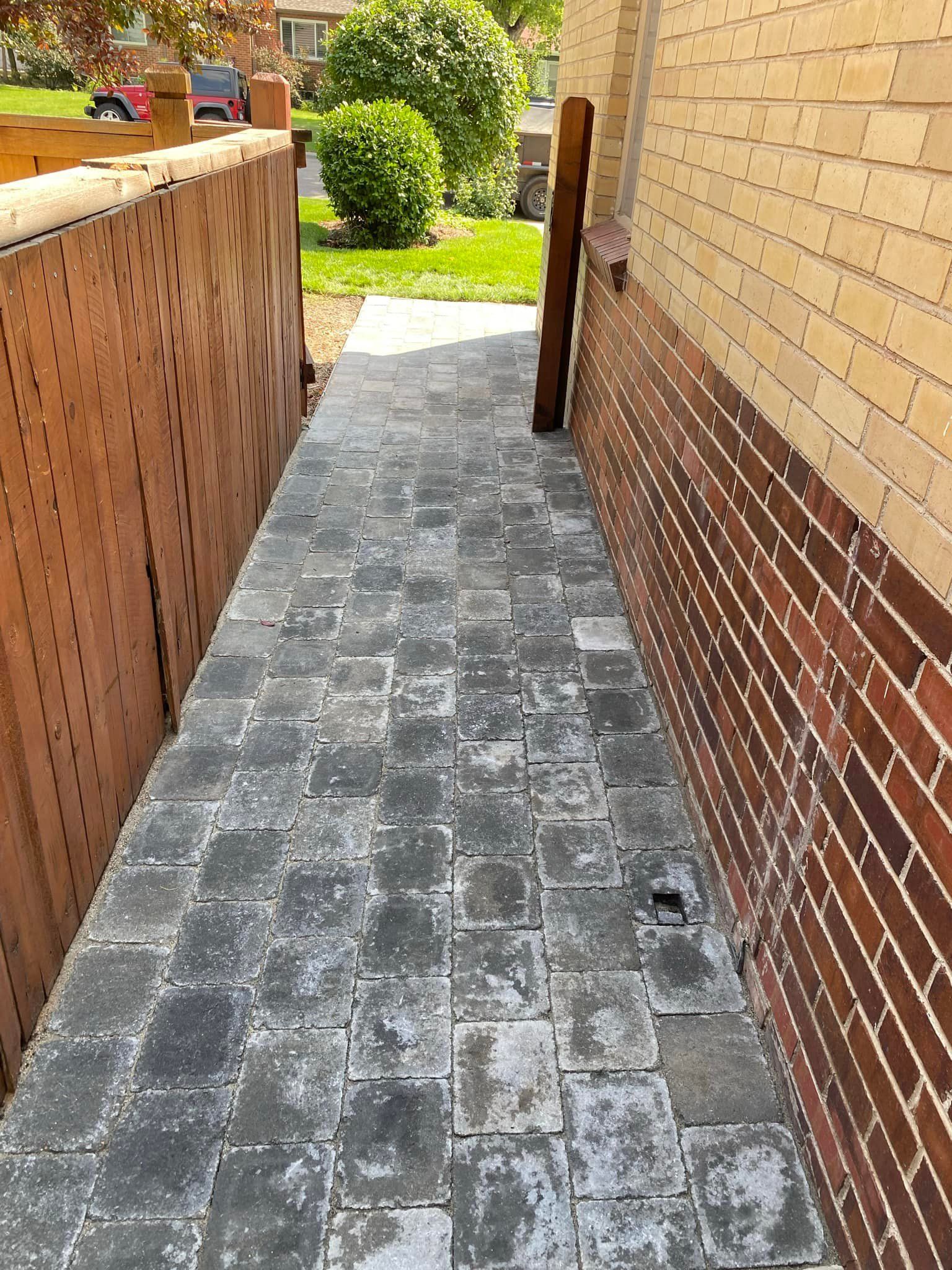 Narrow paved walkway between a wooden fence and a brick wall; green shrubbery visible in the background.