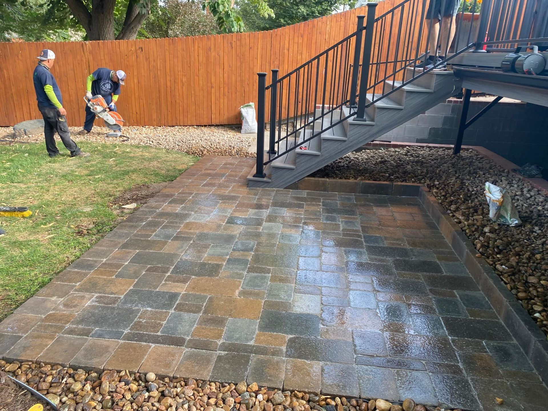 Workers paving a patio with stone pavers near a wooden fence and stairs. 