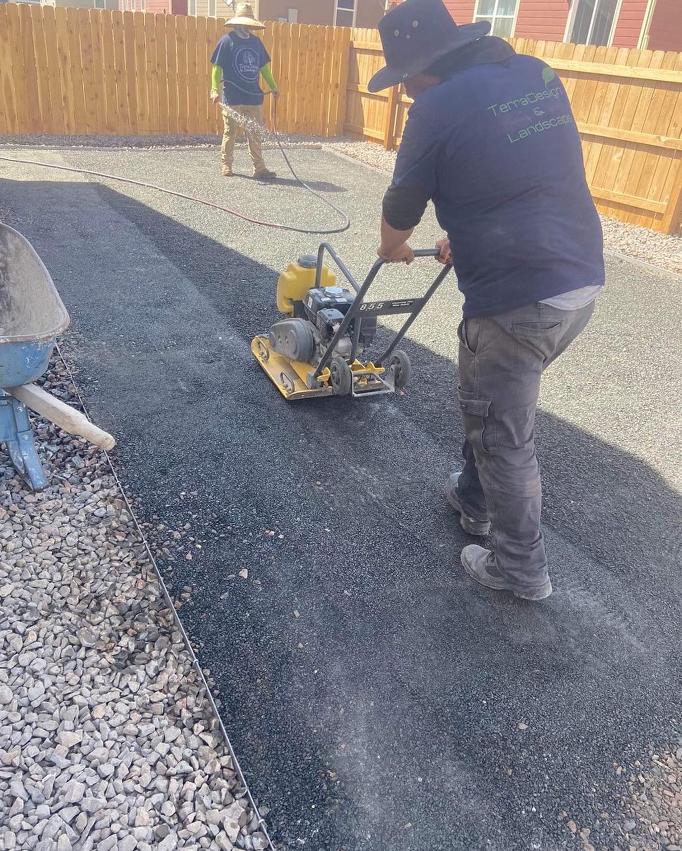 Man operating a plate compactor on a driveway, while another worker rakes gravel in the background. 