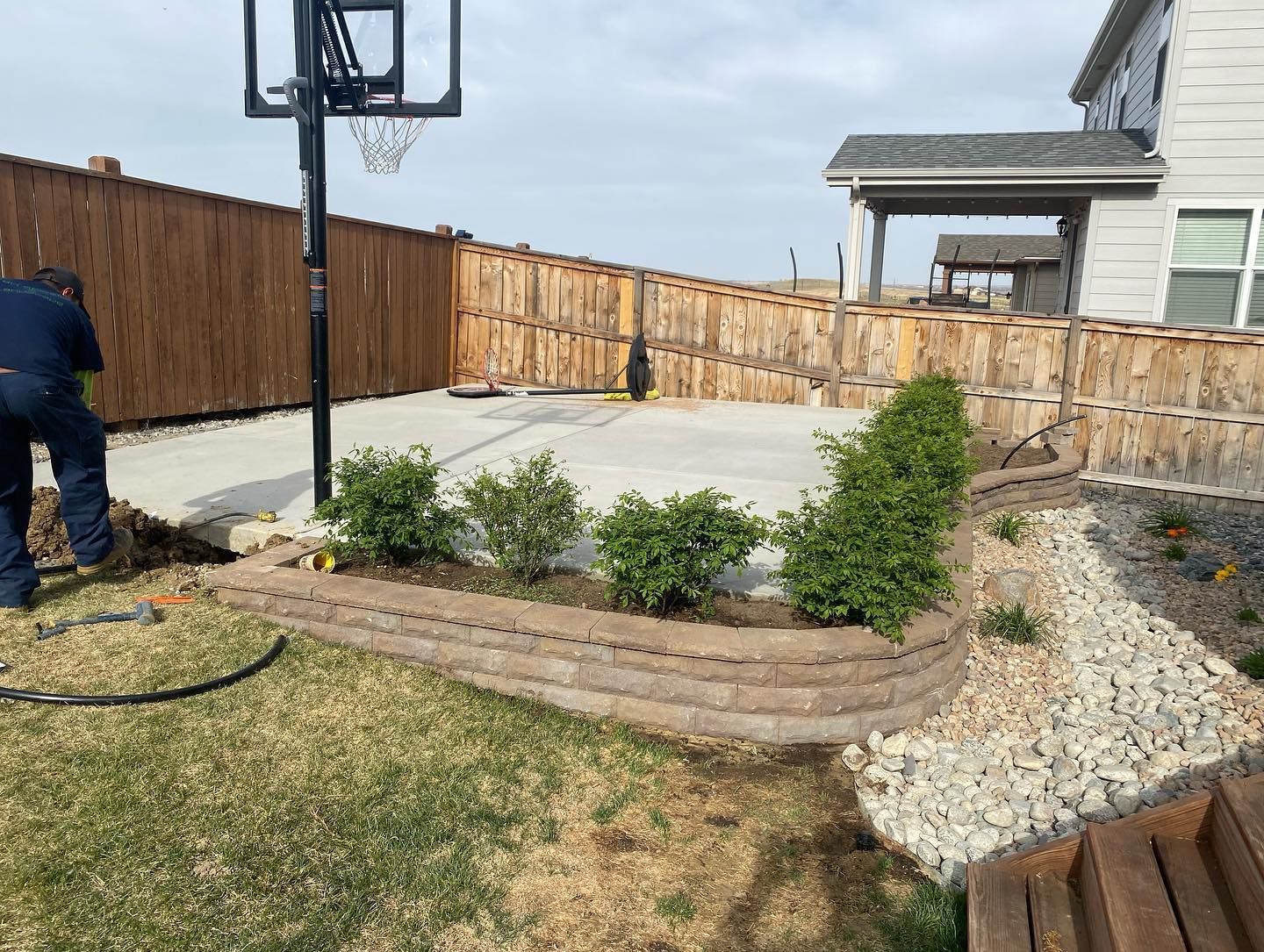 Backyard with concrete patio, retaining wall, and shrubs. Person in blue working on the lawn, wooden fence in background.