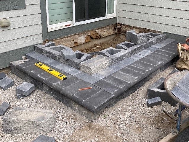 Construction of a gray stone patio and steps outside a house. Blocks and tools are scattered around.