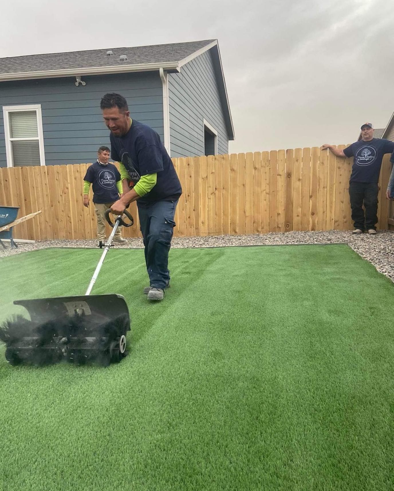 A man uses a machine to groom a patch of green artificial turf in a backyard with two others watching.
