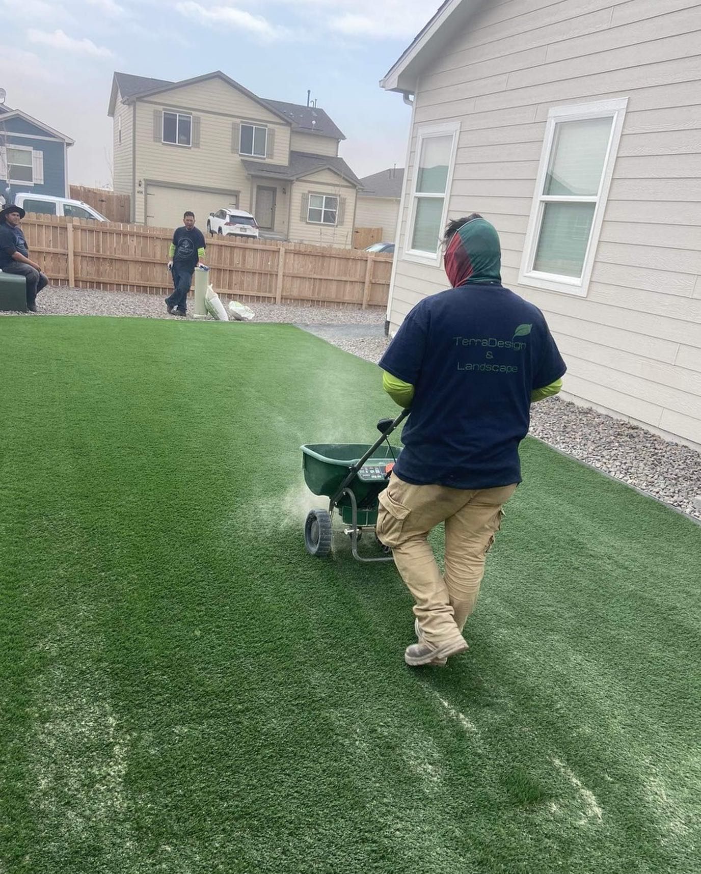 Man spreading material on artificial turf with a spreader. He wears khaki pants, a blue shirt, and a green cap.