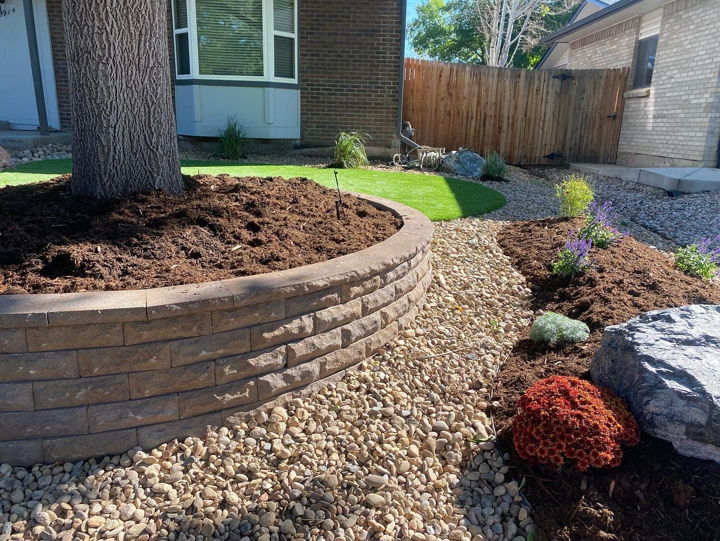 Landscaped front yard with a retaining wall around a tree, rock path, and various plants.