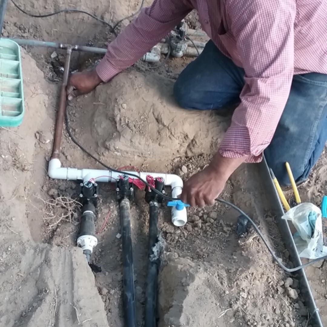 Person kneeling, working on irrigation pipes in a dirt trench. They are connecting or inspecting the system, likely outdoors.