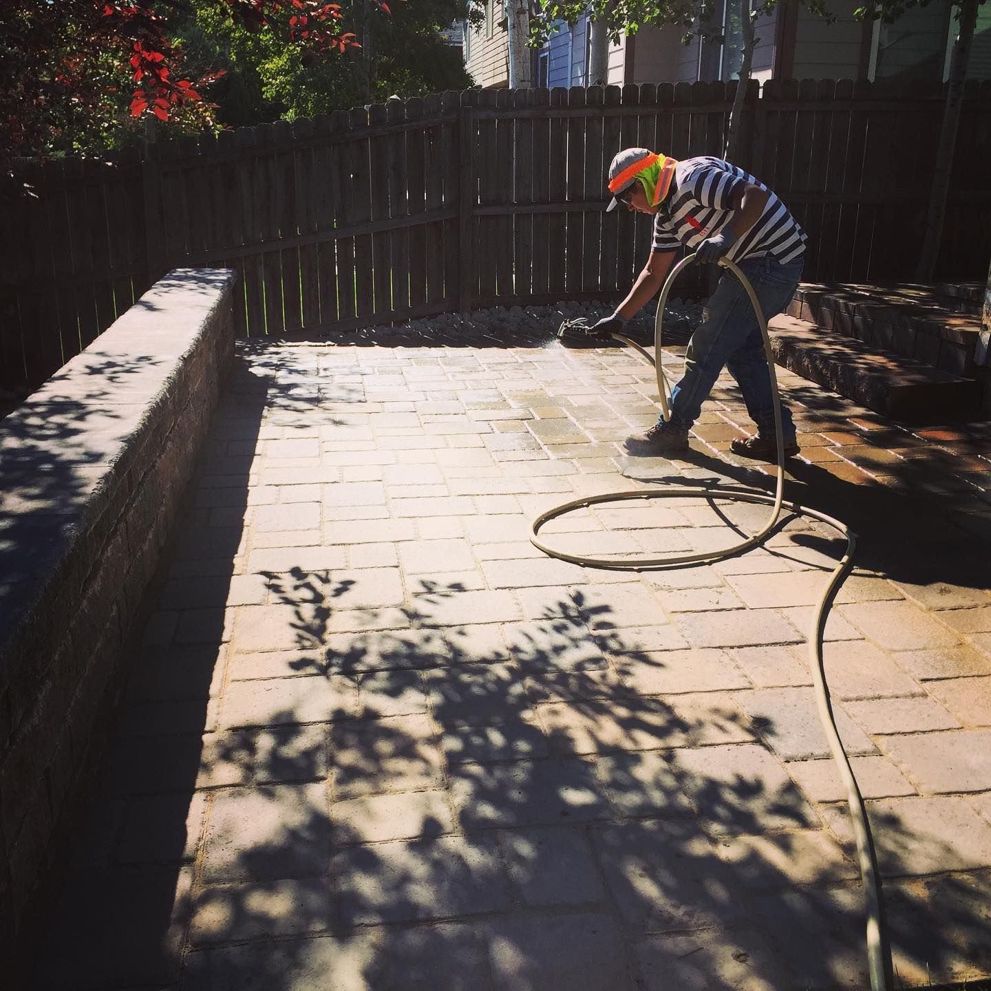 A person pressure washes a brick patio, wearing a striped shirt, safety glasses, and a hard hat. They're in a sunny backyard.