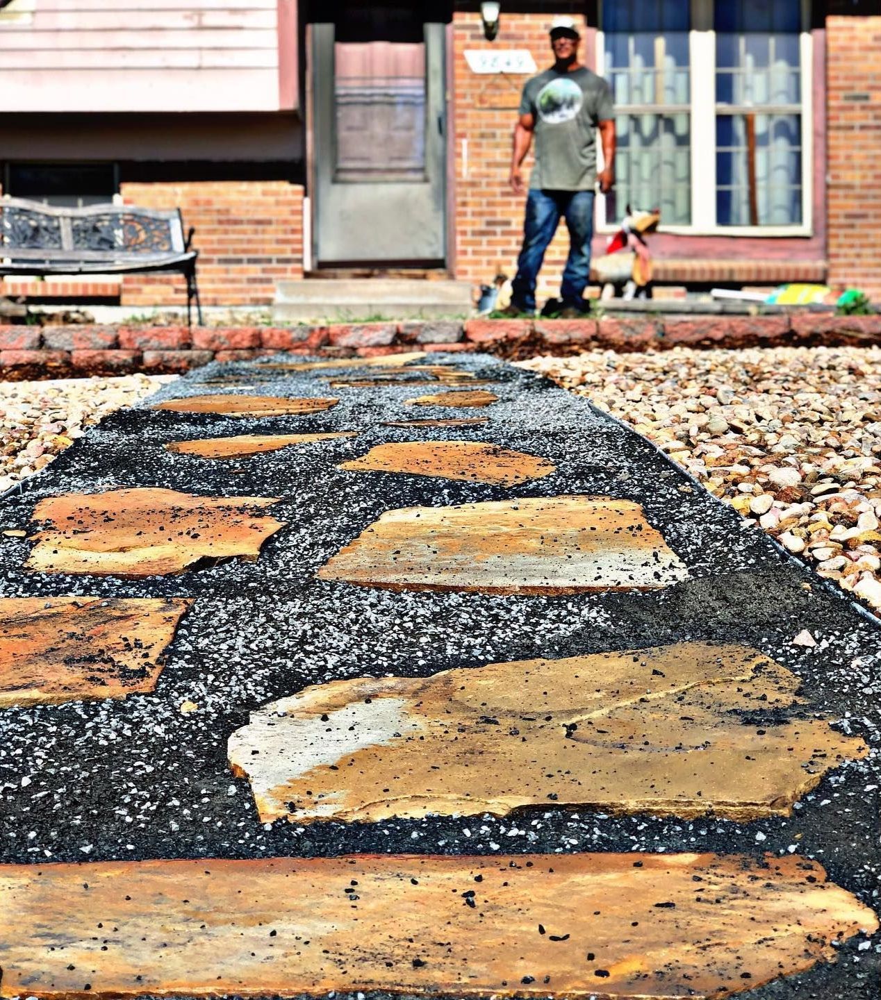 A man standing near the entrance to a brick home with a stone pathway leading to the door. A child is visible near the steps.