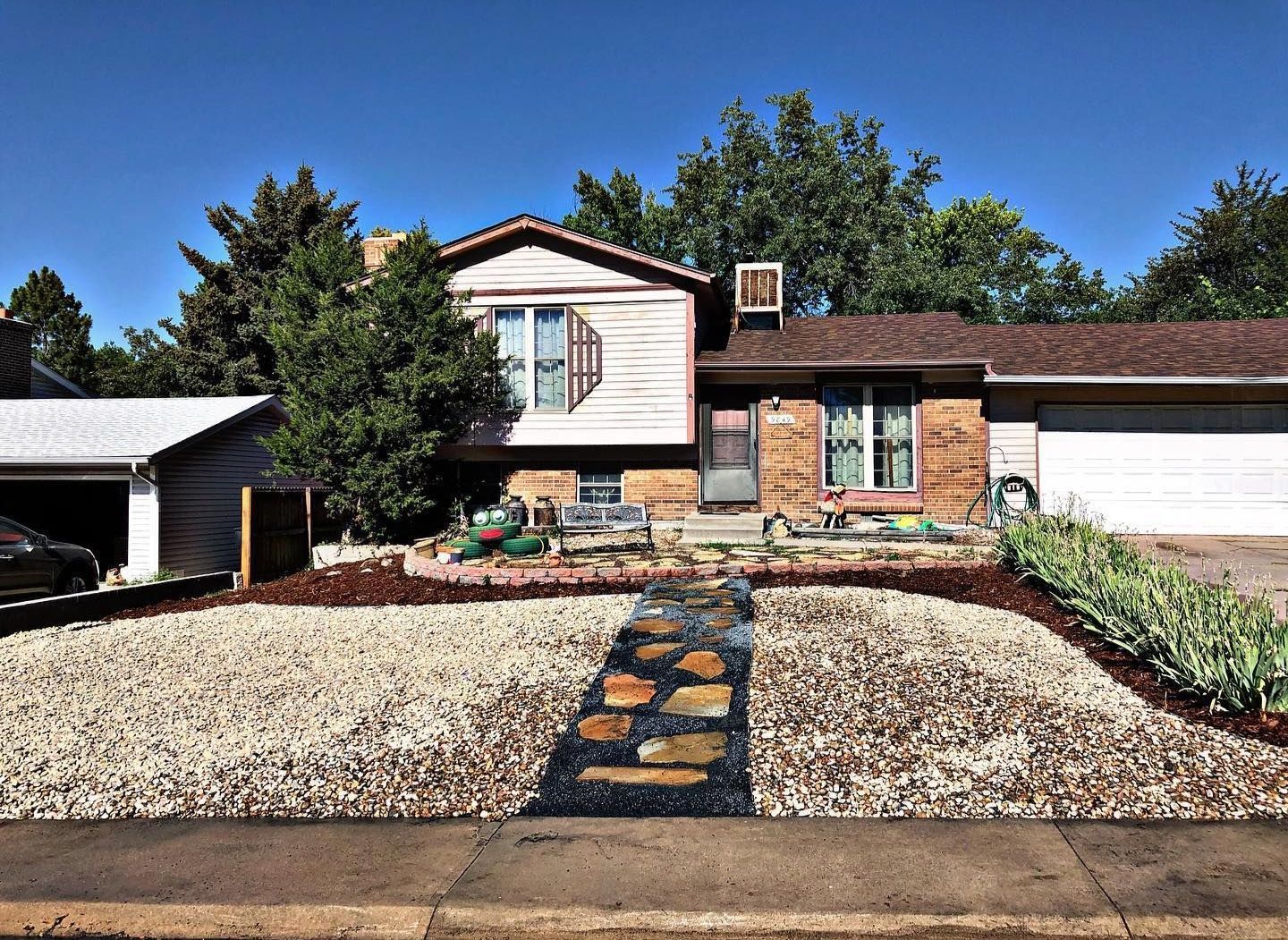 House with beige siding, brown roof, and a rock and mulch landscape in front. A garage and trees are visible.