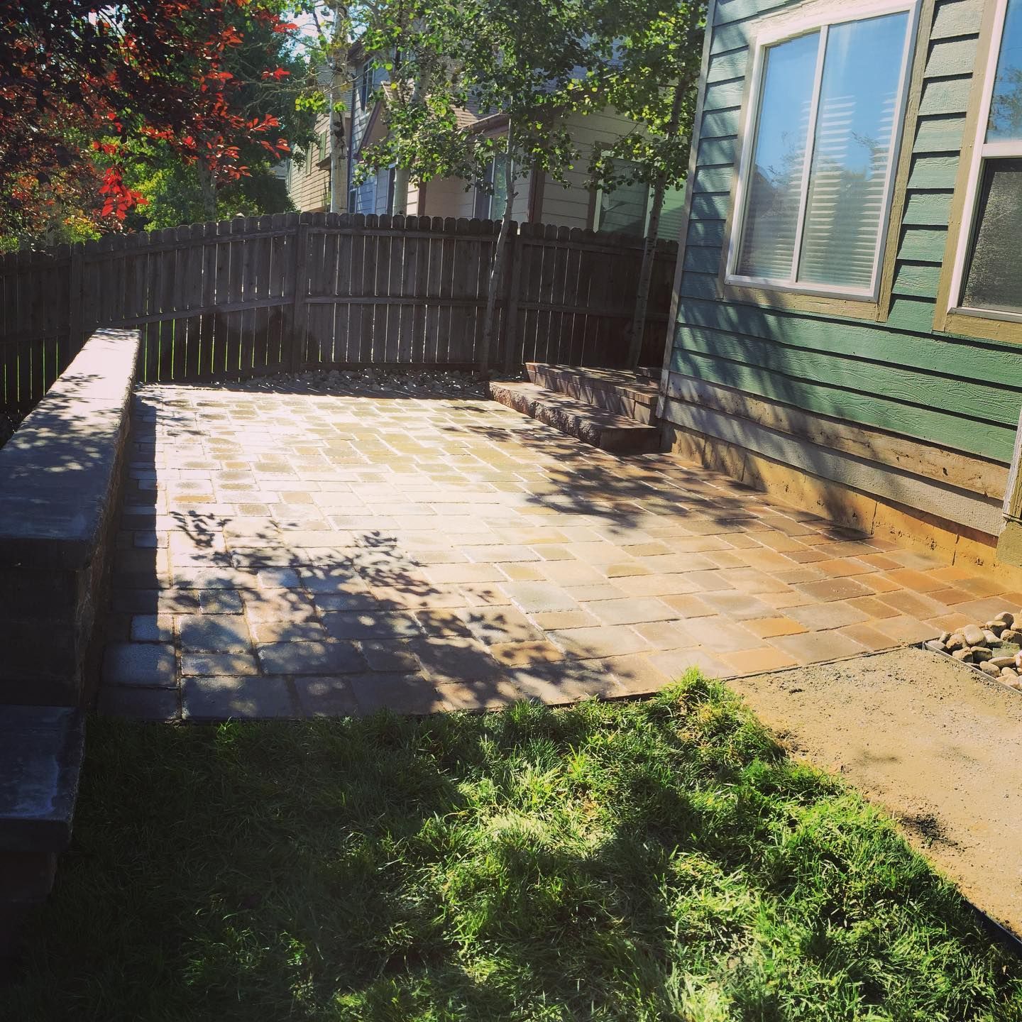 A small backyard patio with brick pavers next to a green house. A wooden fence and trees are in the background.