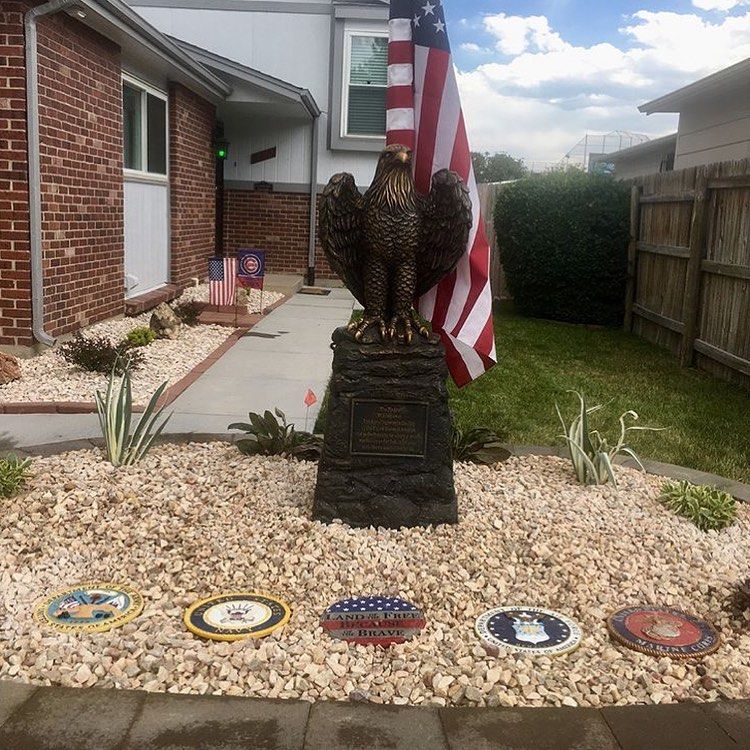 A front yard with a bronze eagle statue, US flag, and military branch plaques on a bed of white rocks in front of a house.