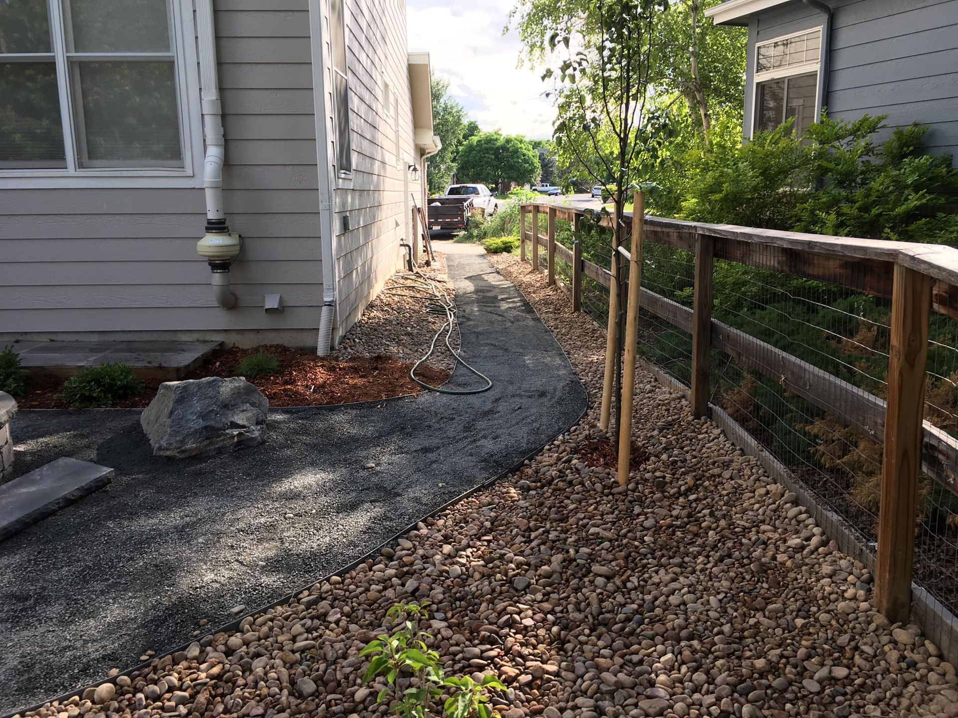 A narrow gravel pathway runs beside a house and wooden fence. Young trees line the path.