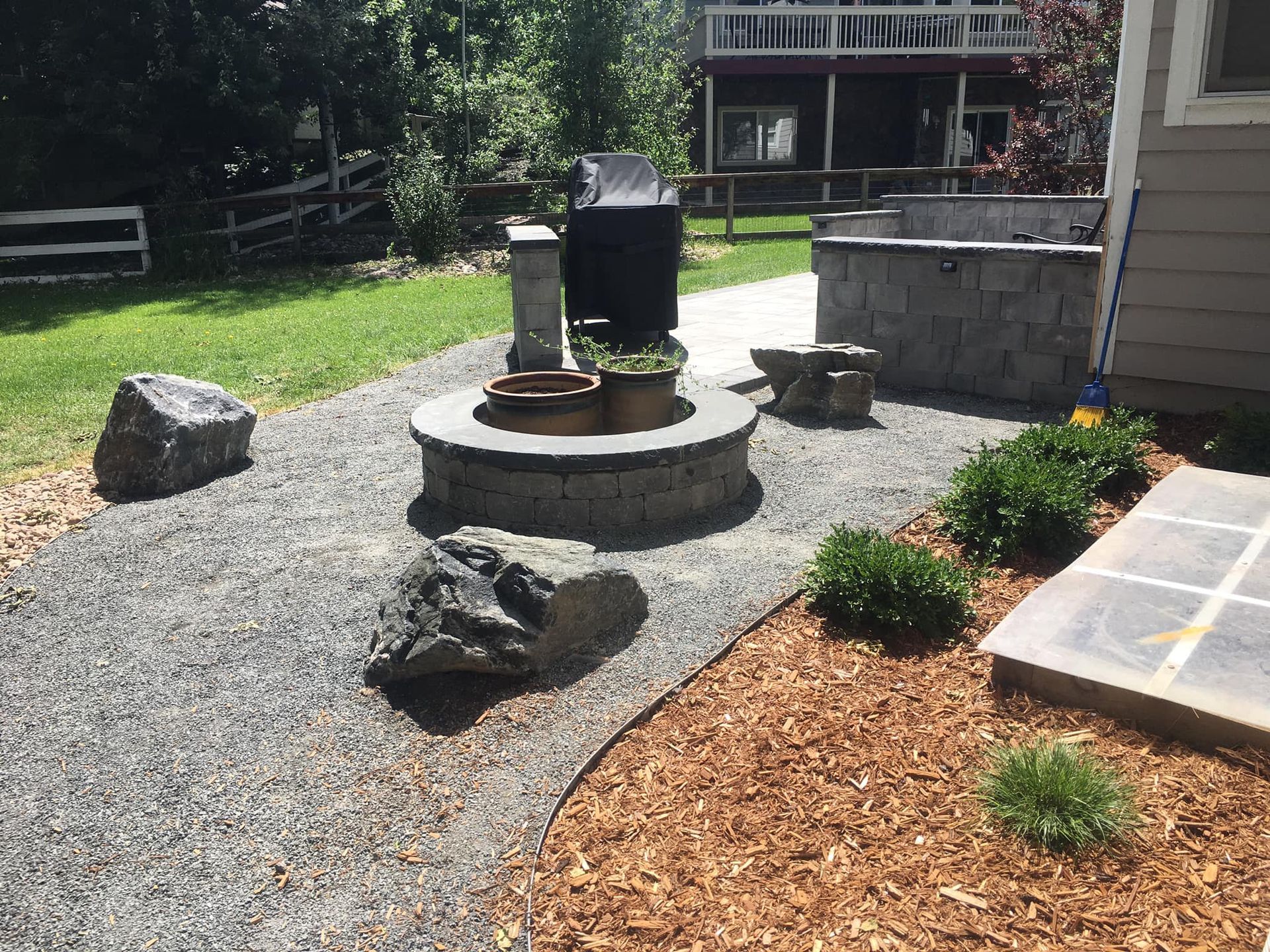 A backyard landscape featuring a fire pit, gravel path, large rocks, and a small seating area.