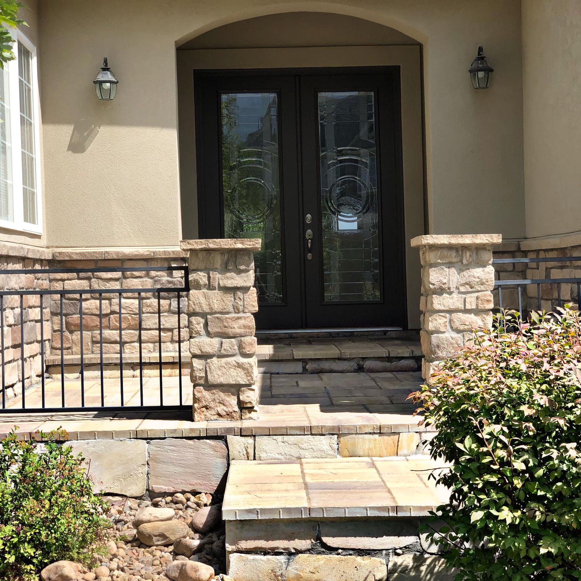Tan house entrance with dark double doors, stone steps, and a black iron railing. Two sconces flank the door.