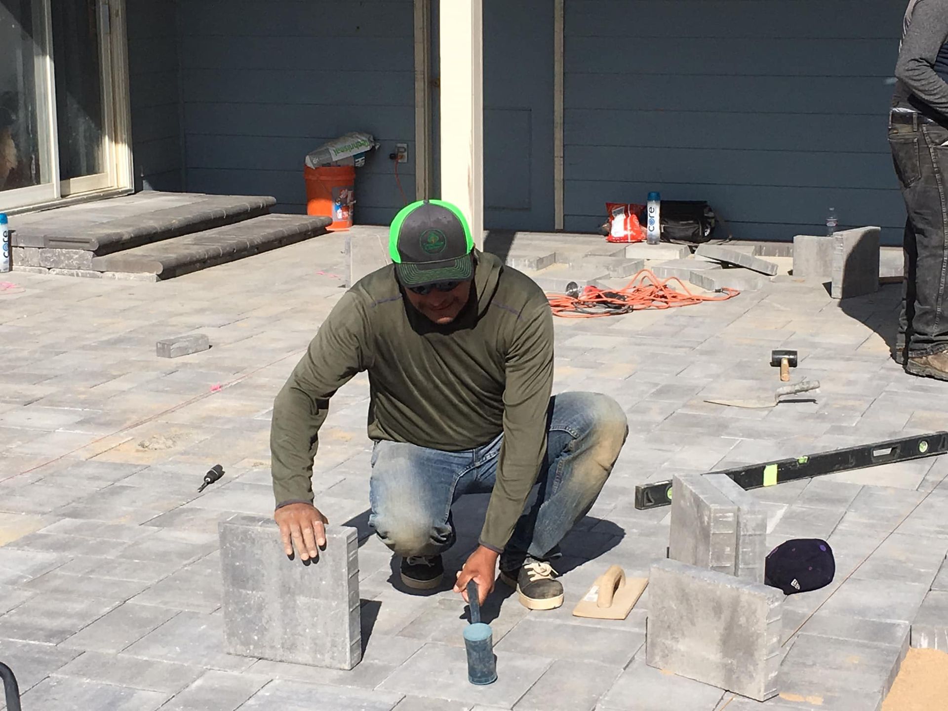A construction worker kneels on a patio, placing pavers, with tools and building materials scattered around.