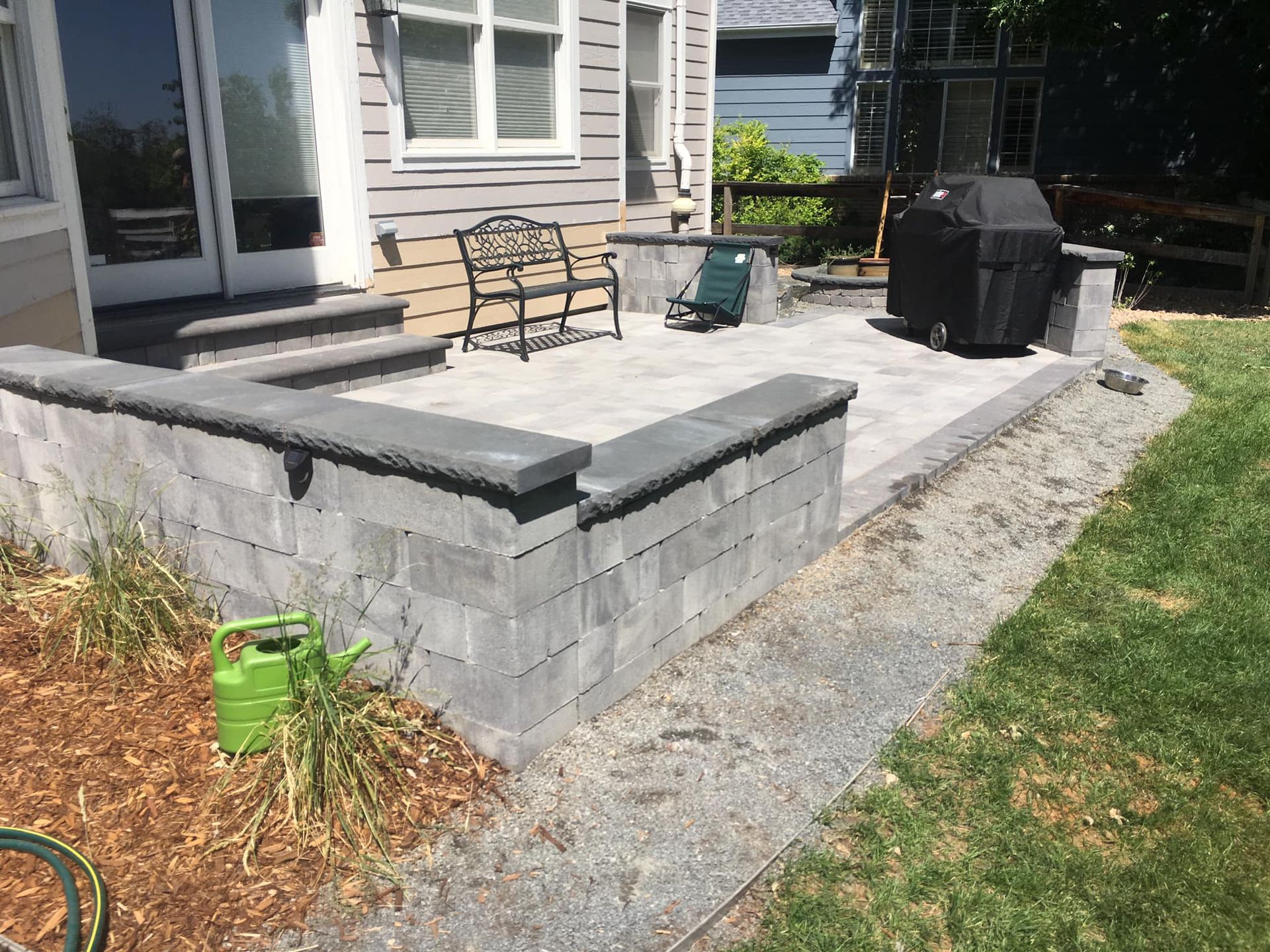 Grey brick patio with raised borders, steps leading to a door, a grill, and a wrought iron bench.