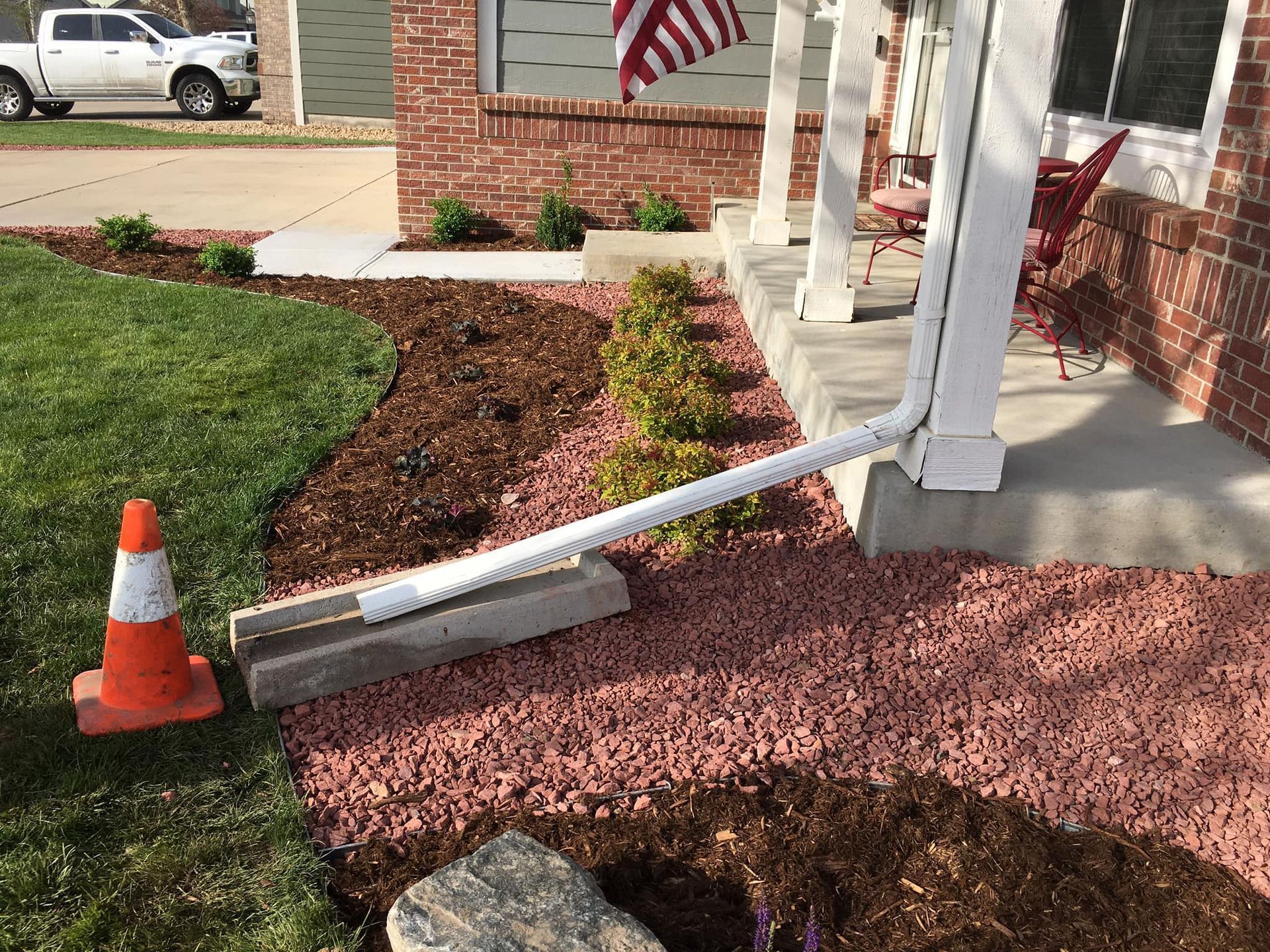 A white downspout rests on concrete blocks, leading to a red gravel bed near a house with a lawn and a safety cone.