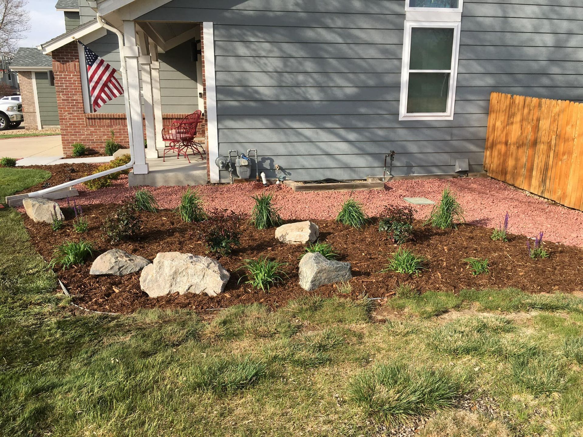 A house with a gray exterior and a red brick porch. A flower bed in front has plants, rocks, and red mulch.