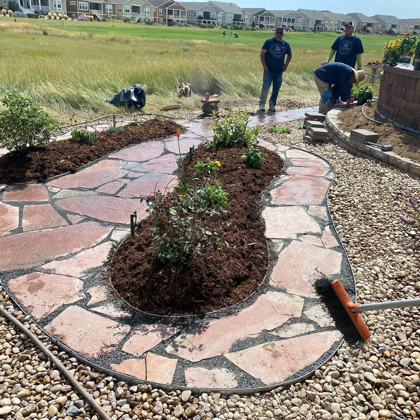People gardening in a backyard with a flagstone path. One person waters plants while others mulch.