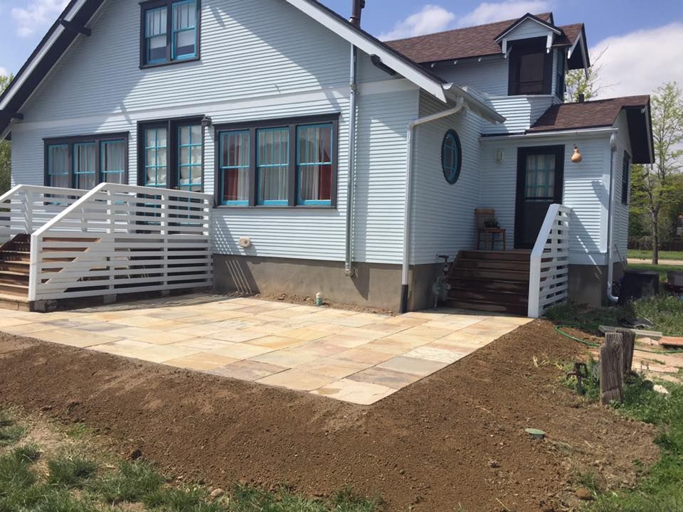 A light blue house with a new stone patio, wooden railings, and brown soil border.