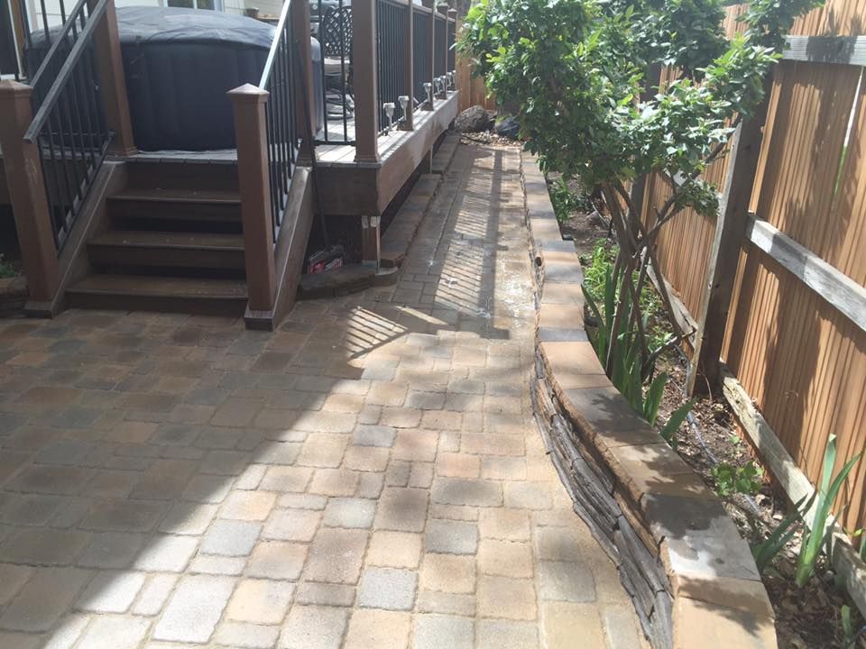 Paver patio with a raised border, leading to a wooden deck with stairs. A wooden fence and greenery are along the side.