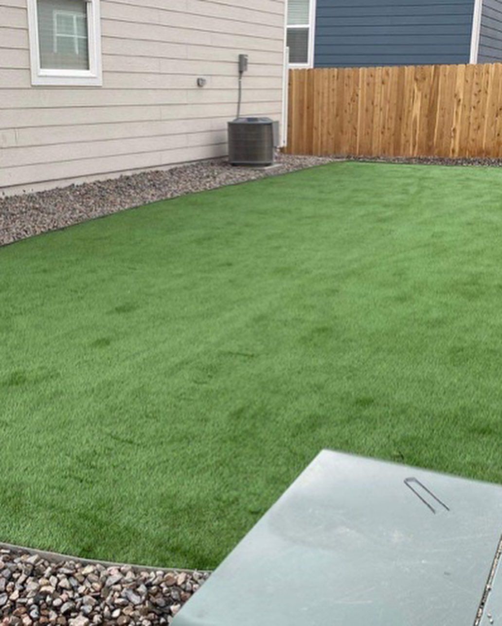 A backyard with artificial green grass, a gravel border, a wooden fence, and a house with a window.