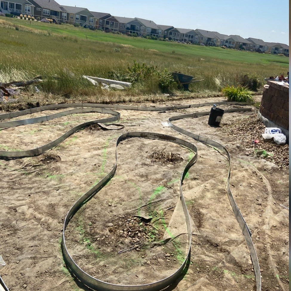 Metal edging outlines a winding path in a dirt yard, with houses on a hillside in the background.