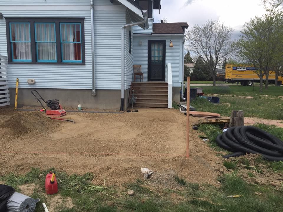A patio under construction next to a light blue house. Gravel is spread on the ground, a compactor sits nearby.