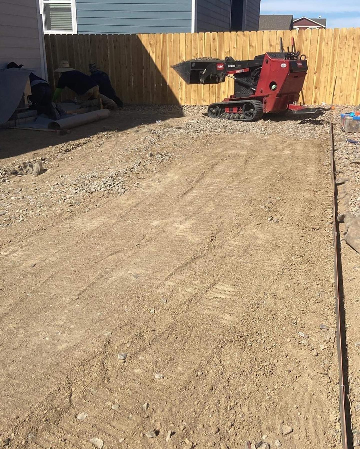 A small construction vehicle working on a gravel and dirt surface in a fenced backyard.