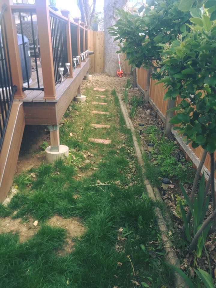 A narrow, grassy side yard with stone stepping stones leading to a wooden fence and a deck with steps on the left.