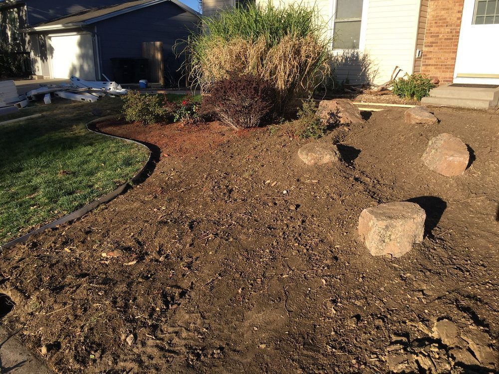 A front yard with freshly tilled brown soil, landscaping rocks, and existing plants near a house. The lawn is green.