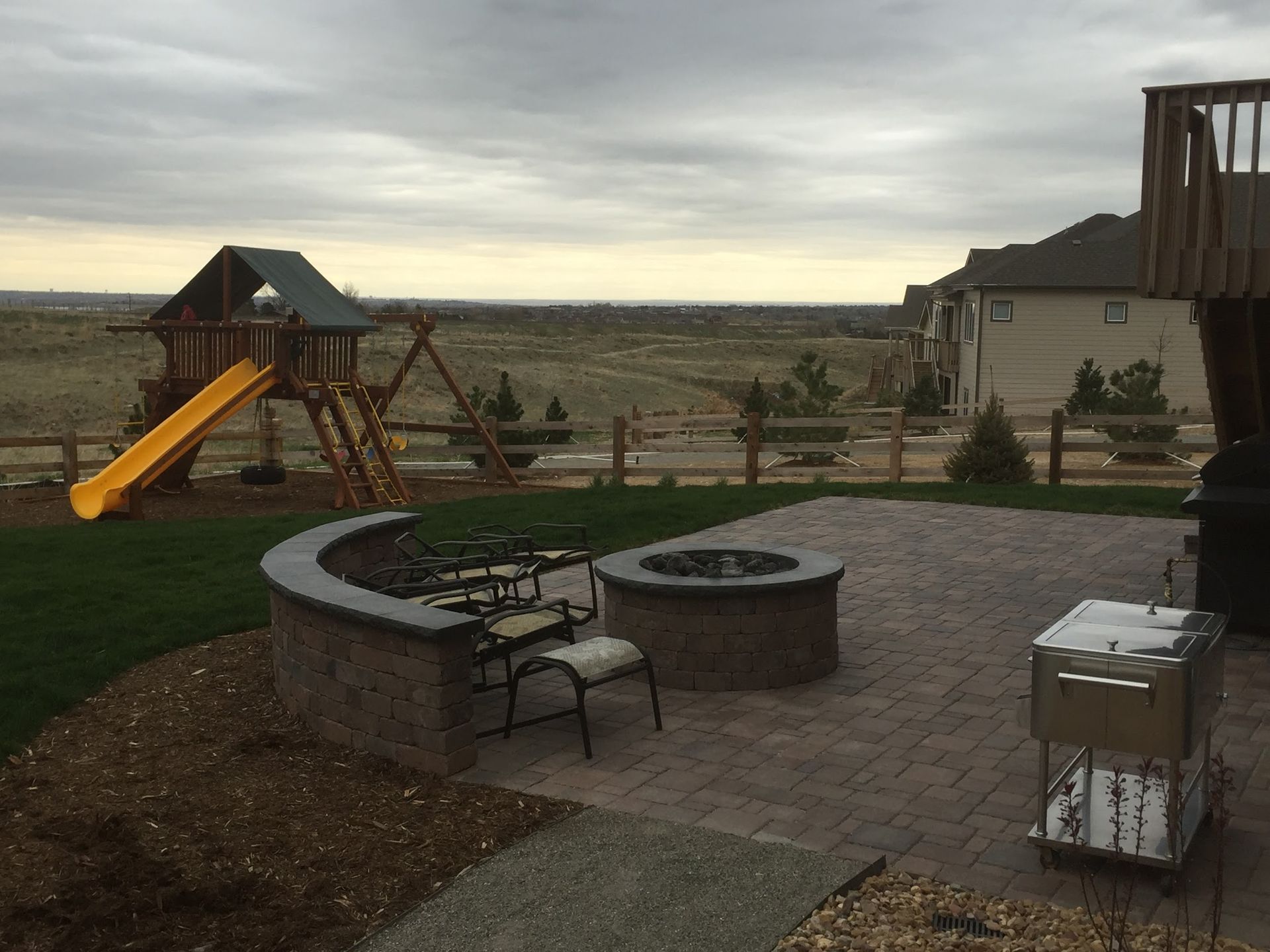 Backyard patio with a fire pit, play set, and a view of a grassy plain under a cloudy sky.