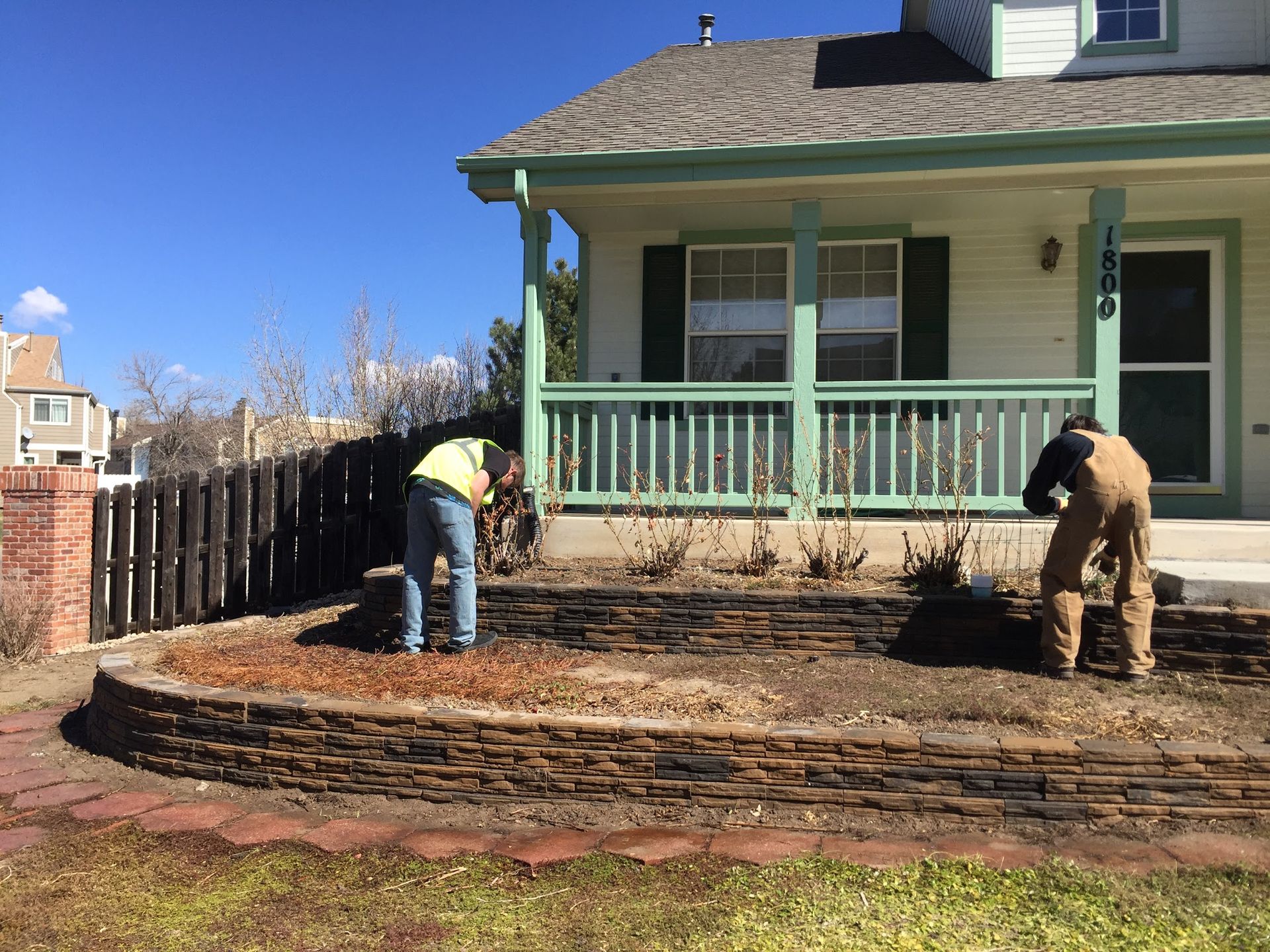 Two workers tending a tiered garden bed in front of a light-colored house with a green porch.