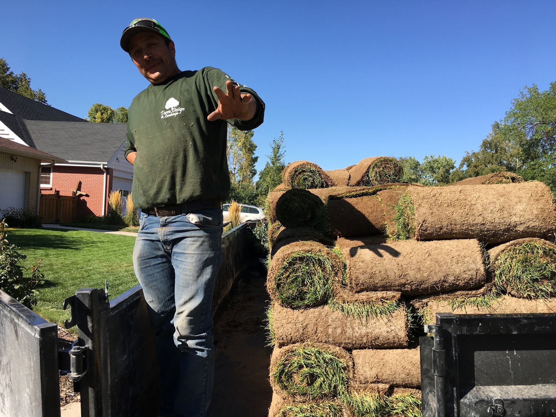 Man standing in a truck filled with rolled-up sod, gesturing with his hand. He's wearing a green shirt, jeans, and a cap.