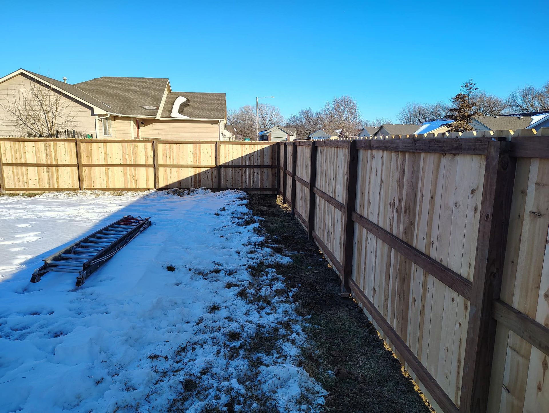 A wooden fence surrounds a snowy yard with a ladder in the foreground.