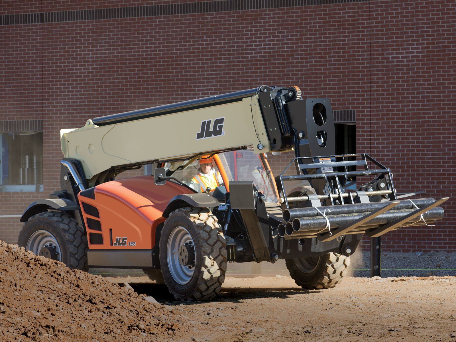 A jlg forklift is driving down a dirt road in front of a brick building.