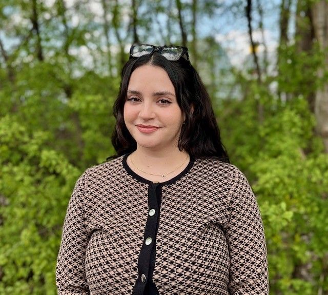 A woman wearing sunglasses and a sweater is standing in front of trees.