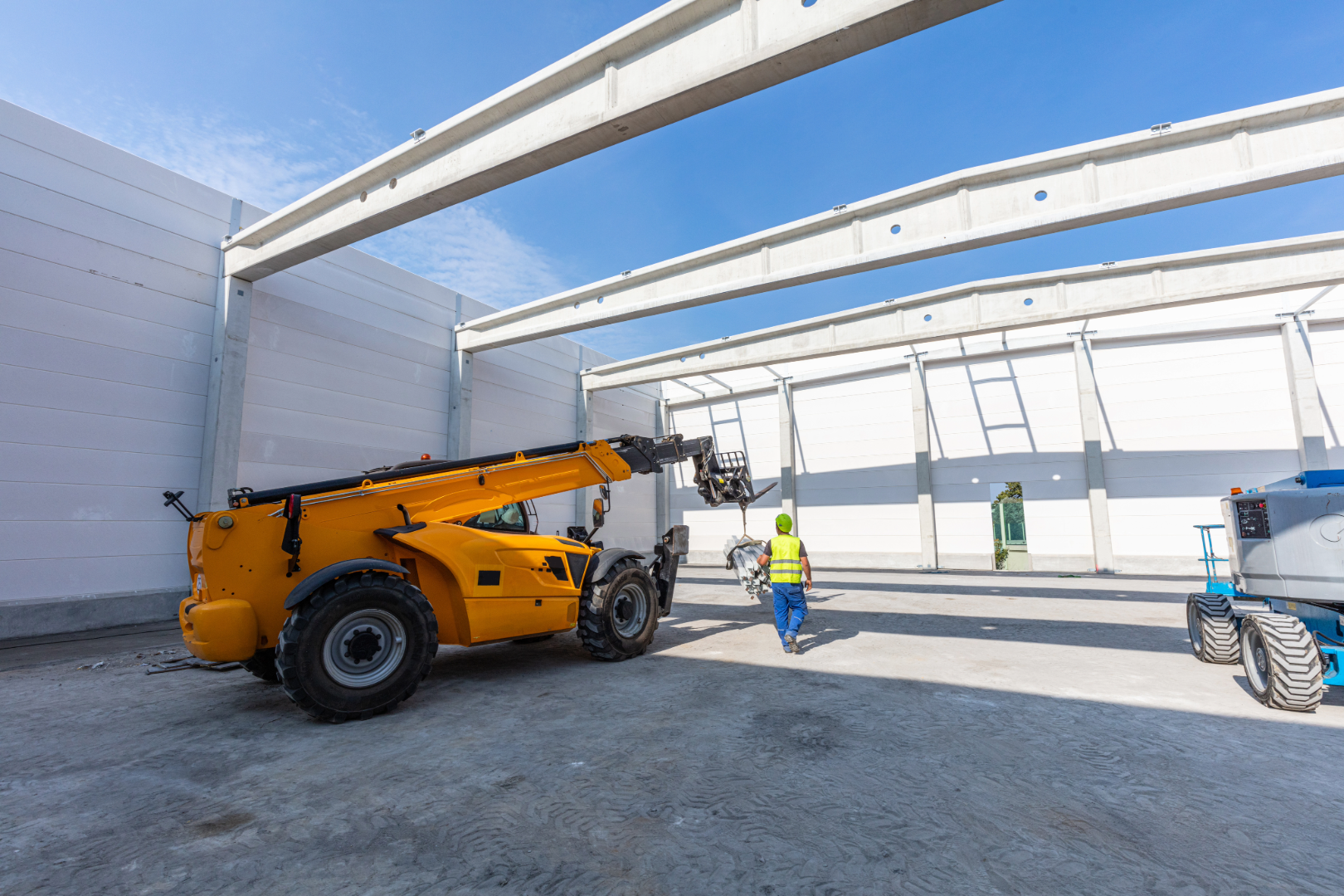 Dieci telehandler lifting material at warehouse build site with worker & Genie boom lift on concrete floor under steel frame
