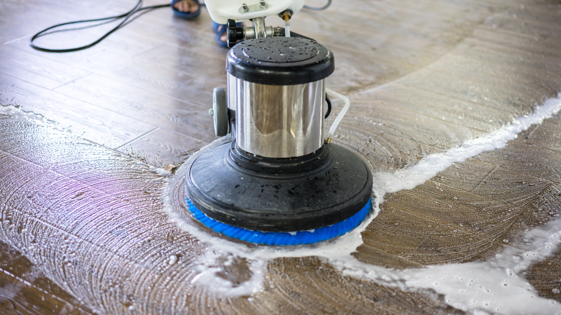 A person in a brown apron and olive pants sweeps a light-colored wood floor in a shop with a bakery display case.