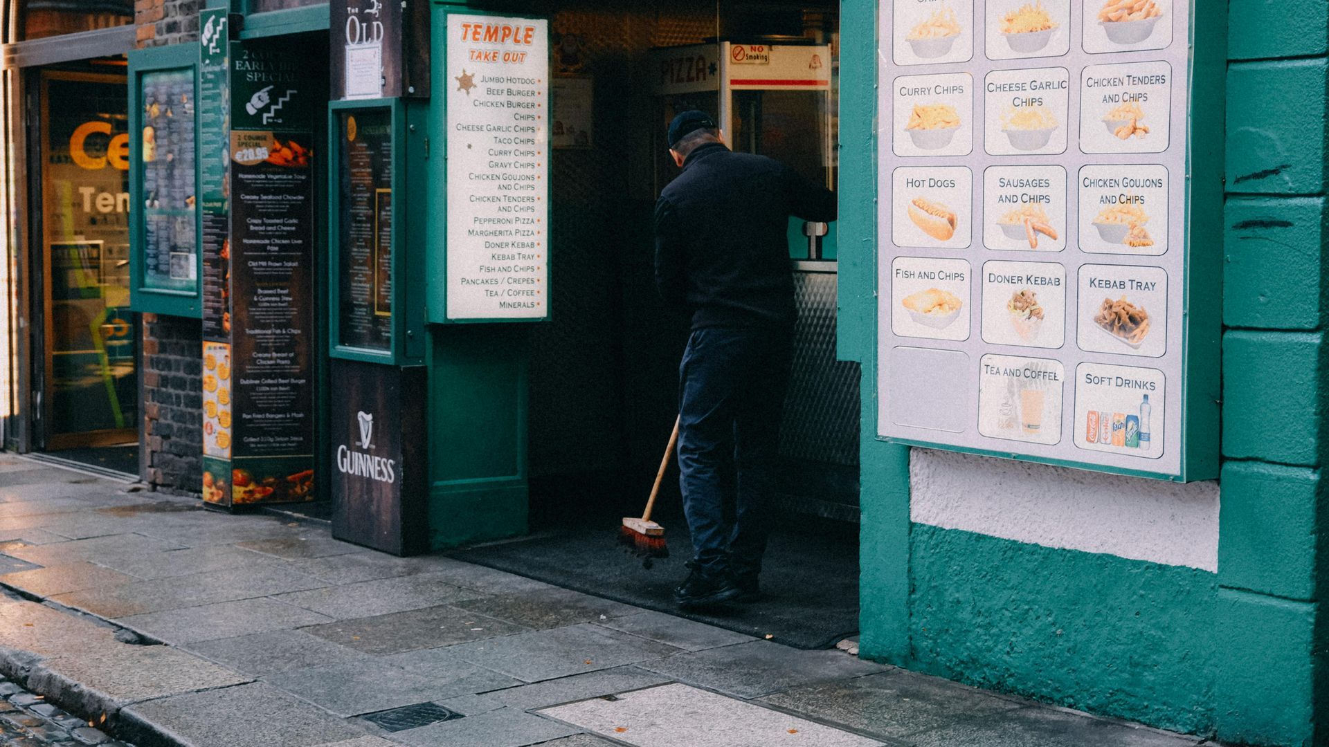 A person in black clothing sweeps the entrance of a storefront with green exterior walls and a food menu displayed.