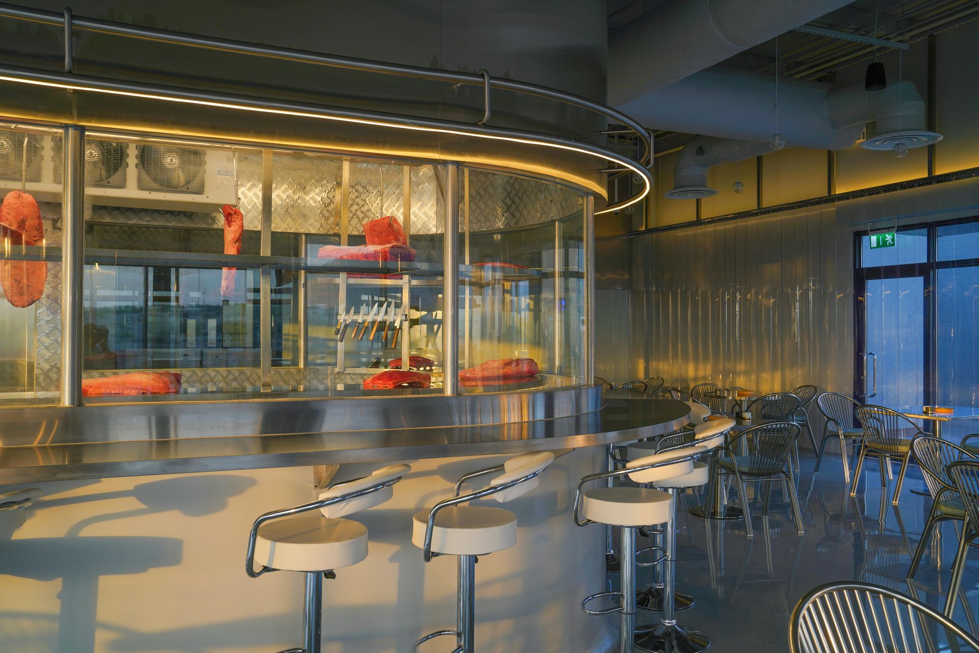 A curved stainless steel bar with white stools in a modern, brightly lit restaurant with hanging cuts of meat.