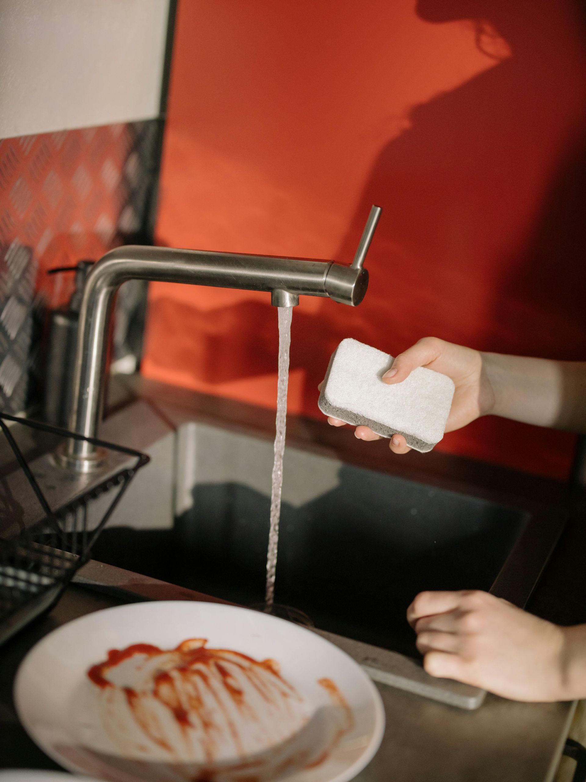 A person holds a white sponge under a running kitchen faucet near a plate with leftover sauce.