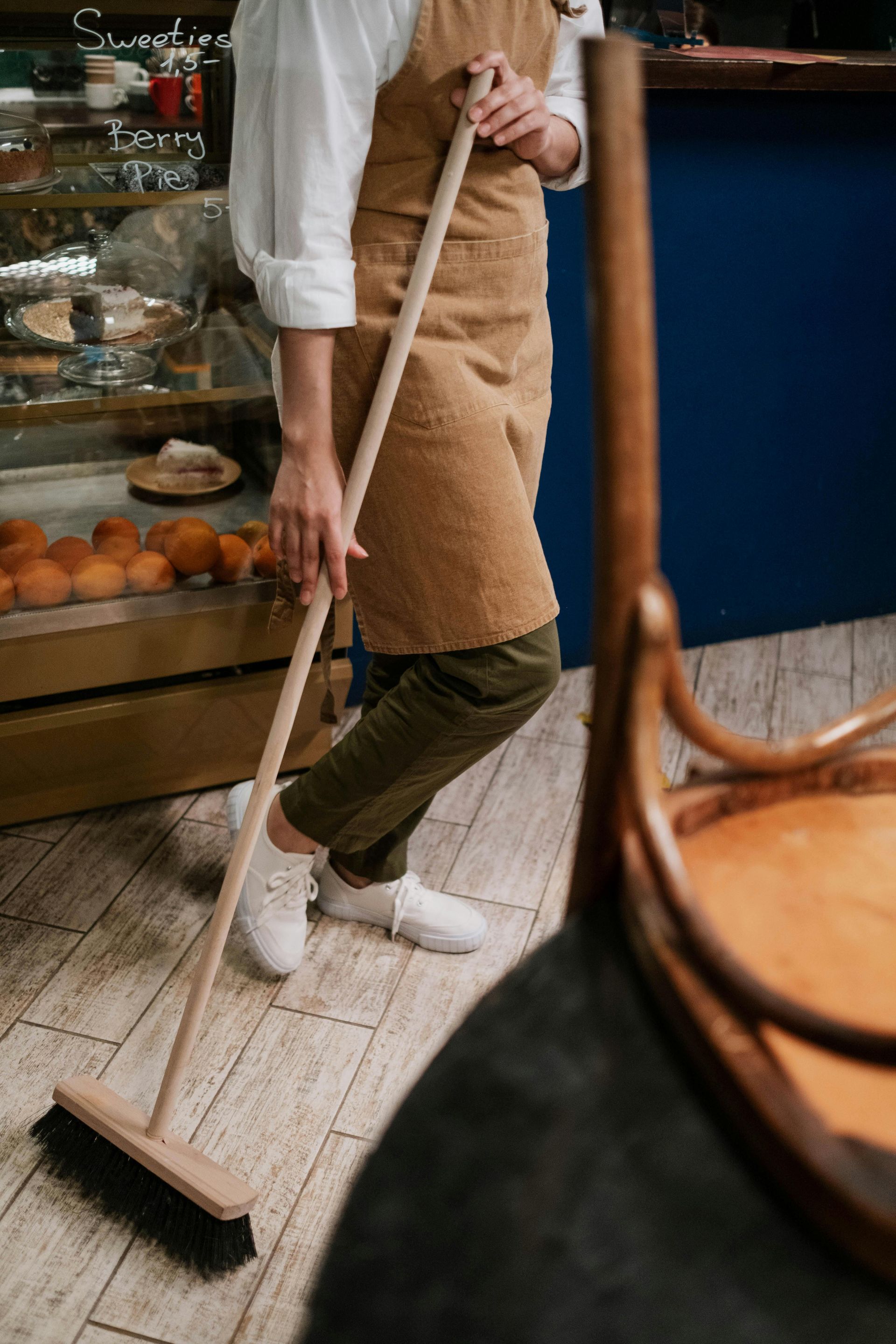 A person in a brown apron and olive pants sweeps a light-colored wood floor in a shop with a bakery display case.